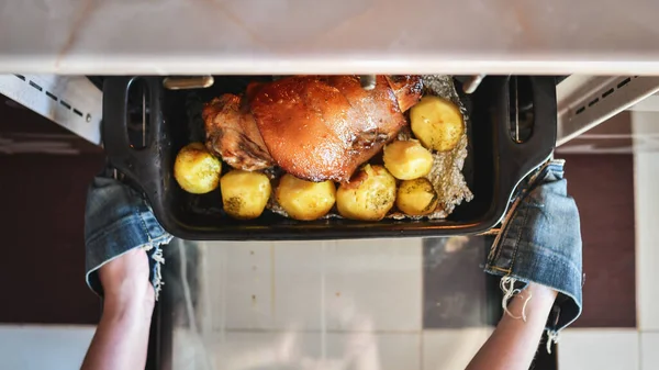 Woman hands taking out roasted pork foreshank with potatoes out of the oven. Top view. Selective focus.