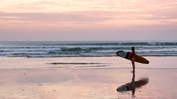 KUTA, INDONÉSIA - JUNHO, 15, 2017: uma menina com um longboard observa o surf na praia de kuta, bali — Vídeo de Stock