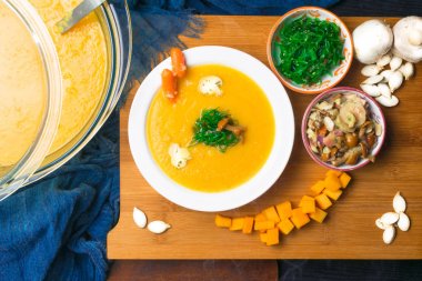 taze sıcak balkabağı çorbası cam pan ve beyaz tabak, ahşap tahta üzerinde mini havuç, kabak tohumu, mantar ve yeşil yosun ile dekore edilmiştir. Vegan çorbası