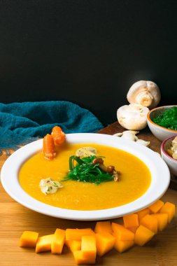 taze sıcak balkabağı çorbası cam pan ve beyaz tabak, ahşap tahta üzerinde mini havuç, kabak tohumu, mantar ve yeşil yosun ile dekore edilmiştir. Vegan çorbası