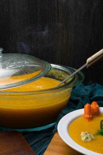 Heiße Kürbissuppe in Glaspfanne, weißer Teller, dekoriert mit Mini-Karotten, Kürbiskernen, Pilzen, grünem Seetang auf Holzbrett — Stockfoto