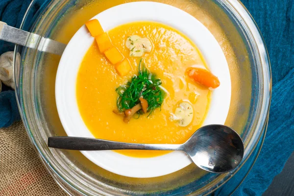 Горячий тыквенный суп в стеклянной сковороде и в белой тарелке, украшенный мини-морковью, тыквенными семенами, грибами, медовым агарическим и зеленым ламинарием на деревянной доске. Веган . — стоковое фото
