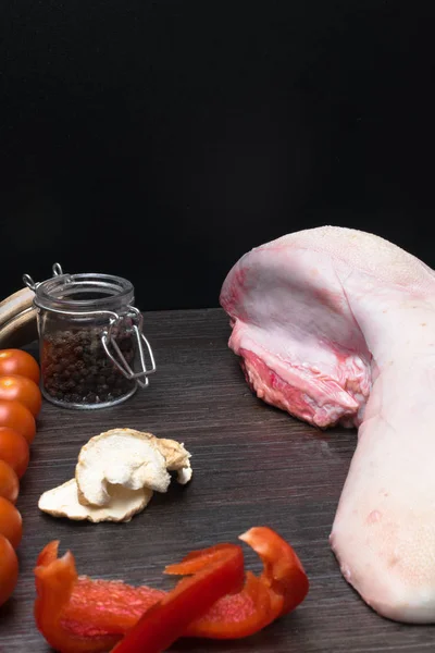 La carne cruda o la lengua de cerdo con hortalizas - el apio, el pimiento negro y rojo, el ajo, la cebolla, yace en la tabla de cortar, sobre el fondo negro el primer plano — Foto de Stock