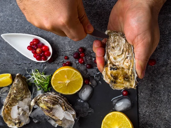 Un uomo che sgusciava ostriche fresche a mani nude sul tavolo di pietra, fette di limone, micro verdi, ghiaccio, mirtilli rossi sul tavolo — Foto Stock
