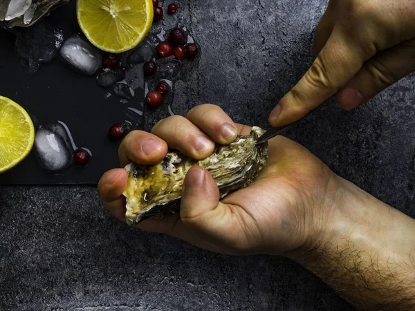 Ein Mann mit einem Messer öffnet eine frische Auster. dunkler Steinhintergrund mit Zitrone, Eis, Beeren, selektiver Fokus. — Stockfoto