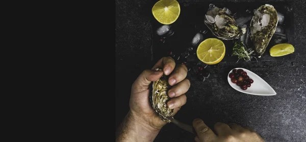 Человек с ножом открывает свежую устрицу. Темно-каменный фон с лемоном, льдом, ягодами, выборочным фокусом . — стоковое фото
