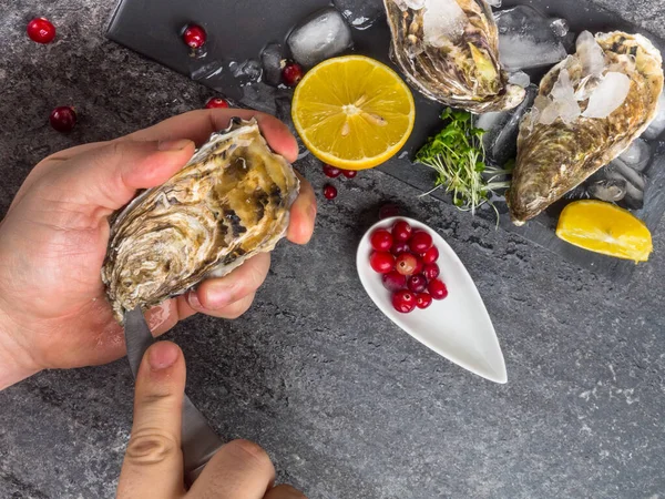 Un uomo con un coltello apre un'ostrica fresca. Fondo in pietra scura con limone, ghiaccio, bacche, focus selettivo . — Foto Stock