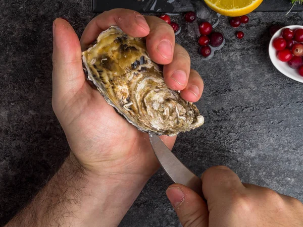 Un uomo con un coltello apre un'ostrica fresca. Fondo in pietra scura con limone, ghiaccio, bacche, focus selettivo . — Foto Stock