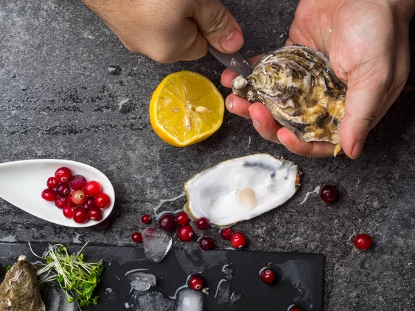 Un uomo con un coltello apre un'ostrica fresca. Fondo in pietra scura con limone, ghiaccio, bacche, focus selettivo . — Foto Stock