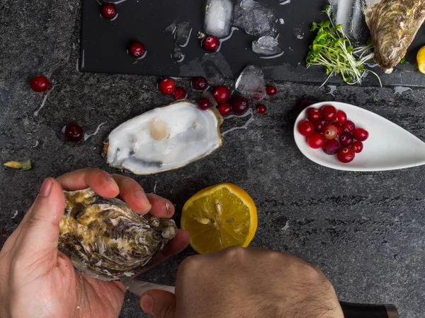 Un hombre con un cuchillo abre una ostra fresca. Fondo de piedra oscura con limón, hielo, bayas, enfoque selectivo . — Foto de Stock
