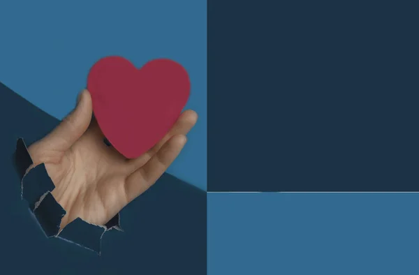 Papel desgarrado azul con agujero, mano sosteniendo el corazón rojo, amor, símbolo de la pasión sobre fondo de color azul de moda para el mensaje — Foto de Stock