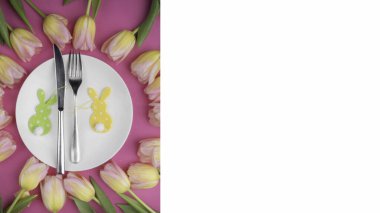 Beyaz tabakta paskalya çatal bıçak seti ya da dekoratif yeşil ve sarı tavşanlı masa seti ve pembe arka plandaki çiçek lalelerinden güzel bir çerçeve. Tatil Dekorasyonu.