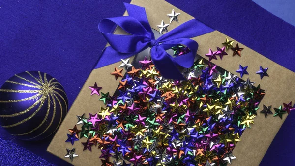 craft gift or present box with satin blue ribbon bow and colorful decorative stars on trendy blue background, Flat lay. Greeting festive card for christmas holiday.