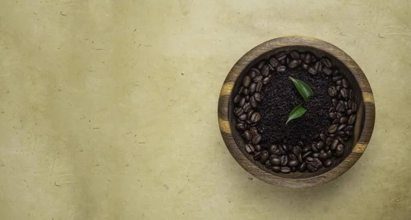 Homemade cosmetics with coffee scrub, beans, and green leaves on wooden bowl on yellow eco friendly background, copy space, flat lay