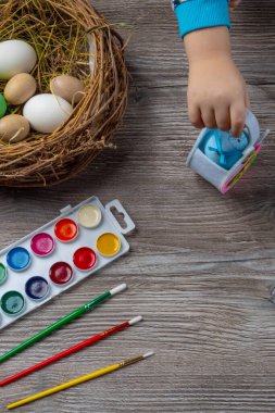 Yuvada paskalya yumurtaları ve ahşap arka planda renkli boya ve fırçalar. Bebek resim yapmaya başlıyor. Seçici odak