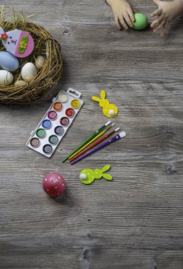 Bebek eli paskalya yumurtasına döner. Ahşap masa boyaları, fırçalar, tavşan, yuva. Yumurta boyamaya hazırlanıyor.