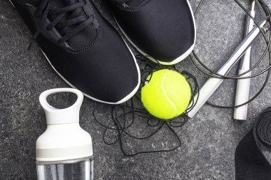 Spor malzemeleriyle kompozisyon, beton arka planda siyah spor ayakkabılar, su şişesi, tenis topu, atlama ipi, boks için el bandajı