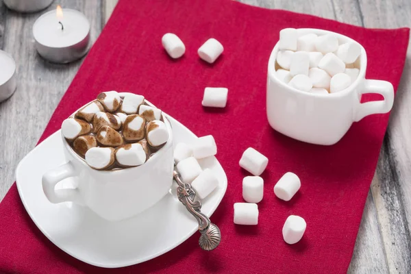 Горячий шоколад с зефиром в белой чашке и две свечи на красном полотенце на деревянном столе — стоковое фото