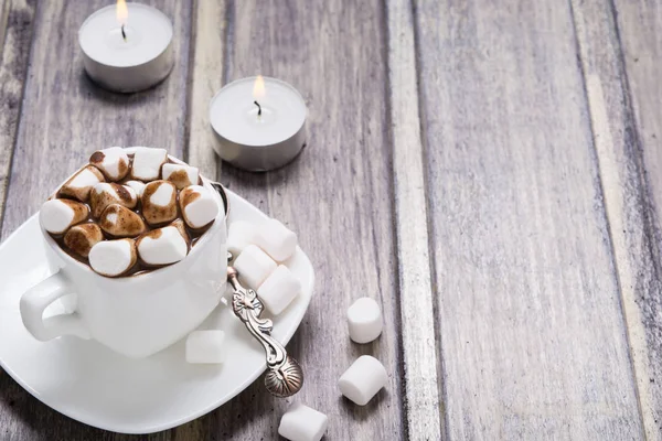 Горячий шоколад с зефиром в белой чашке и двумя свечами на деревянном столе — стоковое фото
