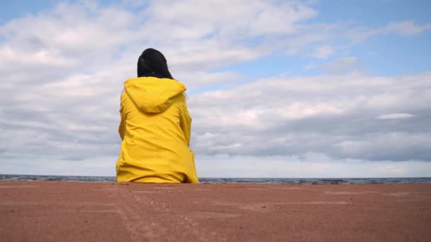 Frau mit dunklen Haaren in gelbem Regenmantel und Jeans sitzt am Strand und schaut auf das Meer und nach dem Aufstehen und geht — Stockvideo