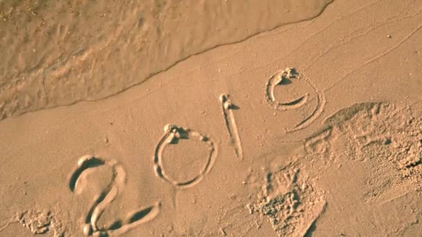 海の波を洗うビーチで砂の上に手書きの2019年のテキスト 新年が来る — ストック動画