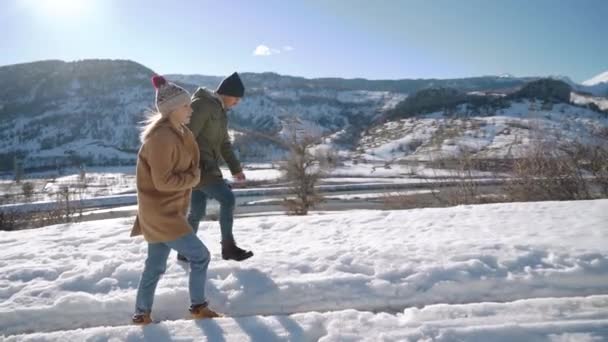 Junges schönes Paar zu Fuß in der verschneiten Straße mit einem Fluss und Bergen im Hintergrund. sonniger Tag. — Stockvideo