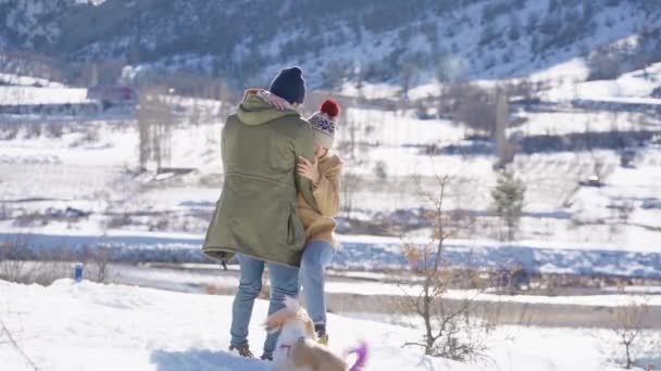 Junges schönes Paar, das sich an einem sonnigen Tag mit einem kleinen aktiven Hund auf einem verschneiten Hügel vergnügt. Winterferienkonzept. — Stockvideo