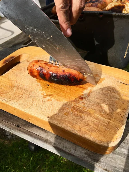 Mano con coltello che taglia una salsiccia su una tavola di legno — Foto Stock