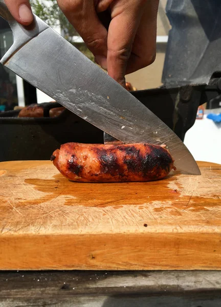 Mano con coltello tagliare una salsiccia su una tavola di legno con griglia sullo sfondo — Foto Stock