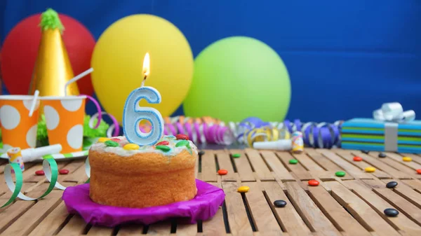 Birthday cake with candles on rustic wooden table with background of colorful balloons, gifts, plastic cups and candies with blue wall in the background. Focus is on cake