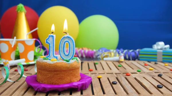 Торт на день рождения со свечами на деревенском деревянном столе на фоне разноцветных воздушных шаров, подарков, пластиковых чашек и конфет с голубой стеной на заднем плане. Фокус на торте — стоковое фото
