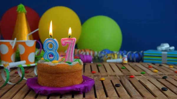 Verjaardagstaart met kaarsjes op rustieke houten tafel met achtergrond van kleurrijke ballonnen, geschenken, plastic bekers en snoepjes met blauwe muur op de achtergrond. Focus ligt op taart — Stockfoto