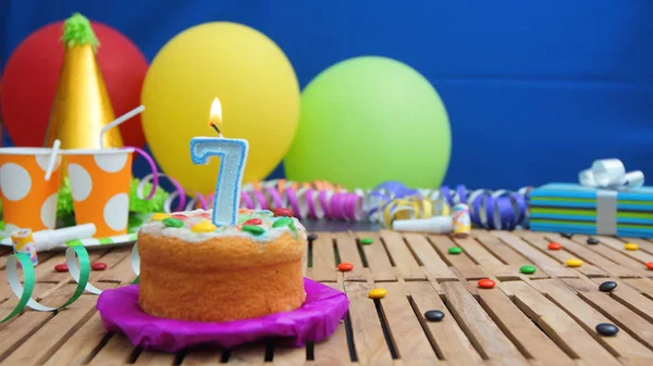 Verjaardagstaart met kaarsjes op rustieke houten tafel met achtergrond van kleurrijke ballonnen, geschenken, plastic bekers en snoepjes met blauwe muur op de achtergrond. Focus ligt op taart — Stockfoto