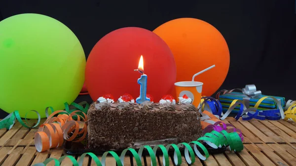 Chocolate birthday cake with candles burning on rustic wooden table with background of colorful balloons, gifts, plastic cups and streamers with black background — Stock Photo, Image