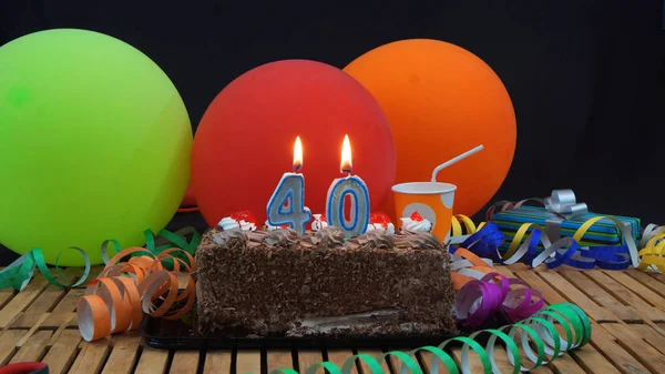 Chocolade verjaardagstaart met kaarsen branden op rustieke houten tafel met achtergrond van kleurrijke ballonnen, geschenken, plastic bekers en wimpels met zwarte achtergrond — Stockfoto