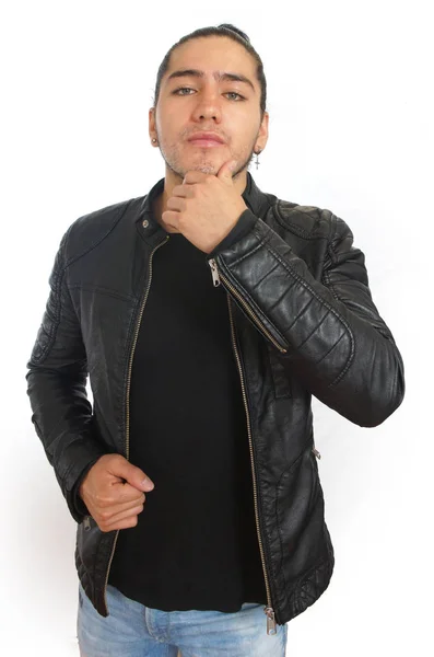 Joven hombre hispano con el pelo recogido arco hecho en camiseta negra y chaqueta de cuero negro, con una mano en la mandíbula y actitud reflexiva, con la mirada hacia la cámara — Foto de Stock