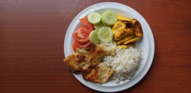 Ev yapımı öğle yemeği tavuk oluşan kabarık beyaz pilav, kızarmış muz, domates dilimleri ve ahşap bir masa üzerinde yuvarlak beyaz tabakta servis salatalık