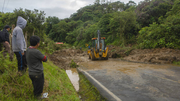 A loader - heavy equipment machine - cleaning a landslide that obstructs the road while people wait to travel the road