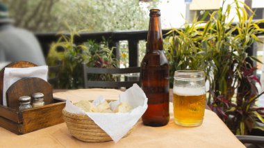 Koyu cam bira şişesi, bira bardağı, ahşap bir masada bir sepet ekmek, odaklanmamış bitkiler.