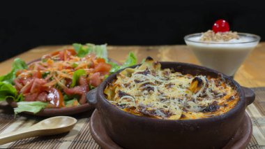 Ahşap bir masa üzerinde havuç, domates ve marul salatası eşliğinde et lazanyalı çamur tabağı.