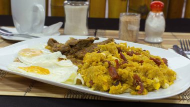 TIGRILO, yanında kızarmış yumurta ve etle birlikte mor bir masada yeşil bir tabakta servis edilen yeşil peynirli muz ve ahşap arka planda bir fincan kahveden oluşan tipik bir yemektir.