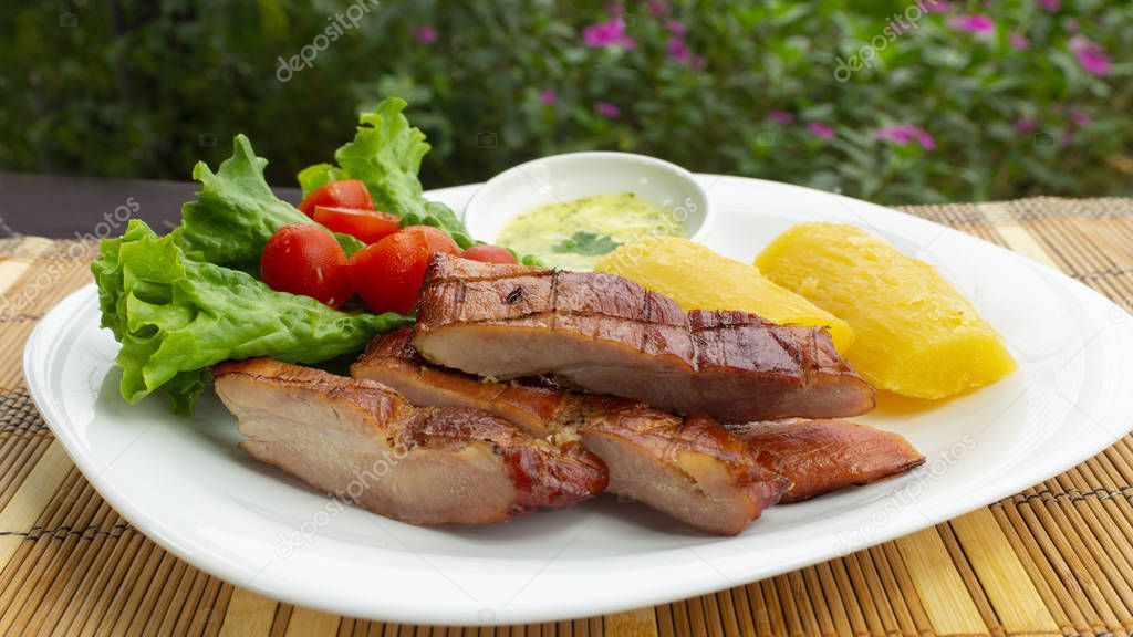White plate with smoked pork meat accompanied by fried yucca and lettuce and tomato salad on wooden table with background of plants and flowers