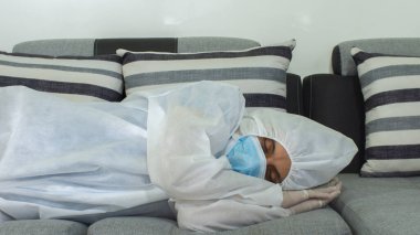 Mavi maskeli ve bioprotatif takım elbiseli doktorun gece beyaz arka planda gri tonlarda büyük bir kanepede uyuduğu ön manzara.