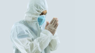 Mavi maskeli ve bioprotatif giysili bir doktorun yan görünüşü. Başı eğik ve gözleri kapalı. Dua ederken ya da meditasyon yaparken bir arada.