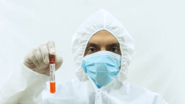 Maskeli doktor ve kolları öne doğru uzamış beyaz bioprotatif giysili doktor ve sağ eli üzerinde kan örneği olan bir tüp ve beyaz arka planda Covid-19 etiketi olan bir test tüpü tutuyor.