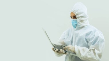 Beyaz bioprotatif takım elbiseli ve mavi maskeli bir doktor ellerinde beyaz arka planda açık bir dizüstü bilgisayarla ayakta duruyor.