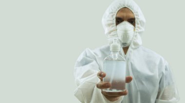Maskeli ve beyaz bioprotatif kıyafetli bir doktor elinde beyaz arka planda bir şişe jel alkol tutuyor.