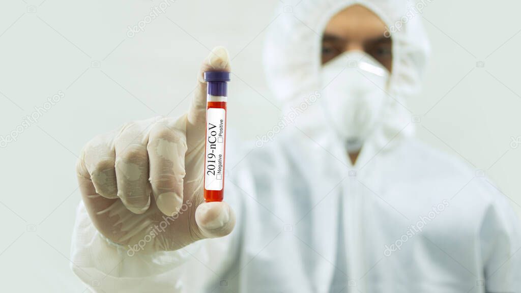 Doctor with mask and white bioprotective suit with arm extended forward and his right hand holding a test tube with blood sample and Covid-19 label in the foreground on white background