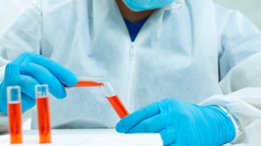 Bioprotatif takım elbiseli, maskeli ve mavi eldivenli Latin doktorların beyaz arka planda iki cam test tüpü arasında kırmızı bir sıvıyı karıştırmasını yakından izleyin.