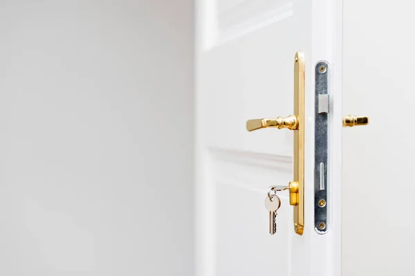 Anahtarlar anahtar deliğinde, beyaz kapının üzerinde altın kapı tokmağı var.